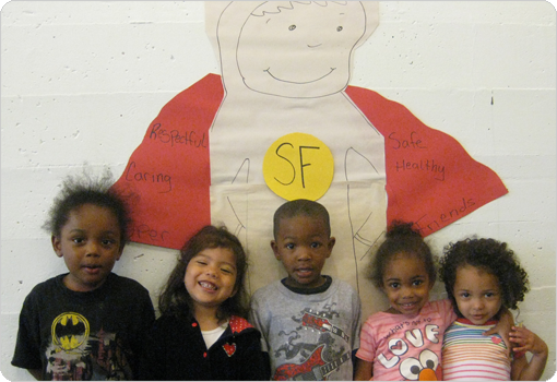 Group of diverse children standing in front of a Super Friend poster